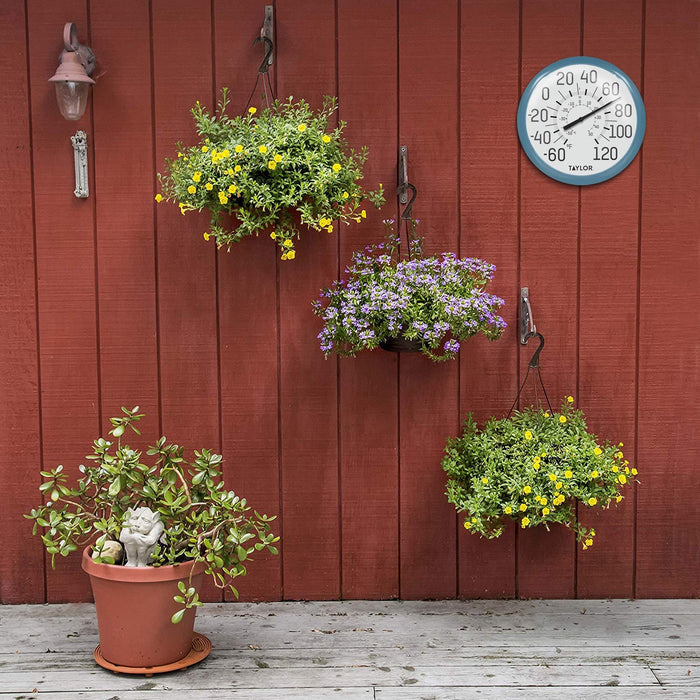 Outdoor thermometer with blue rim mounted on a red wooden fence beside hanging yellow and purple flower baskets.