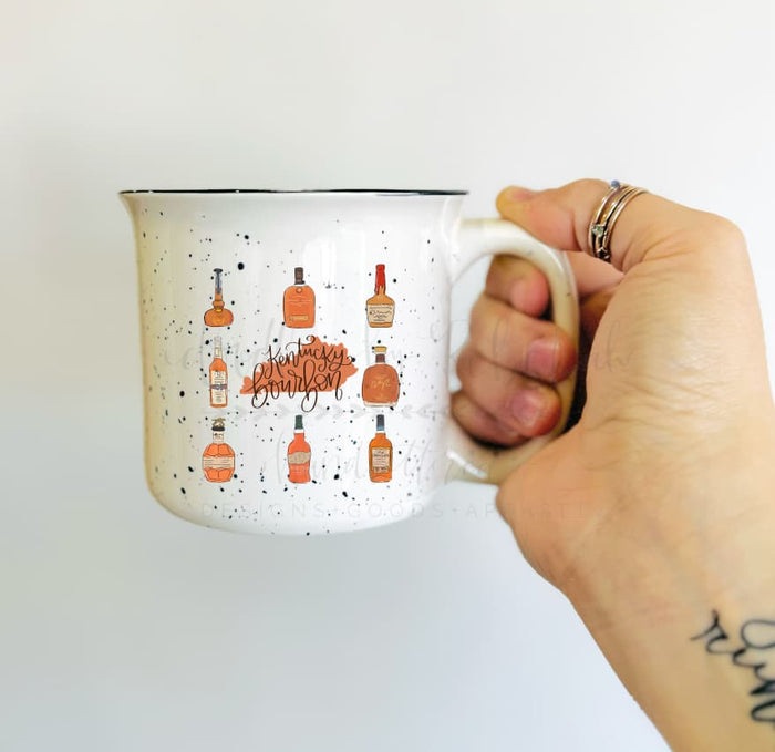 A hand holding a white ceramic mug with brown speckles and an illustration of bourbon bottles and the state of Kentucky