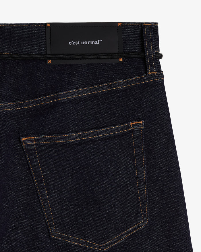 A detailed close-up of the back waist of The Jeans Slim Fit in Rinse Wash, highlighting the black rubber patch with the c'est normal logo, the back pocket, and subtle orange accent stitching.