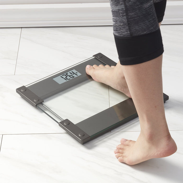 Foot on a modern digital glass bathroom scale with a dark metal frame on a light tiled floor.