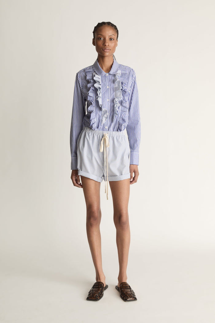 Person wearing a blue and white striped shirt with ruffled details and white shorts on a plain background