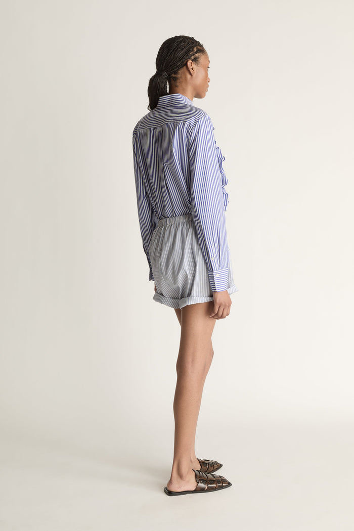 Person wearing a blue and white striped shirt with ruffled details and white shorts on a plain background