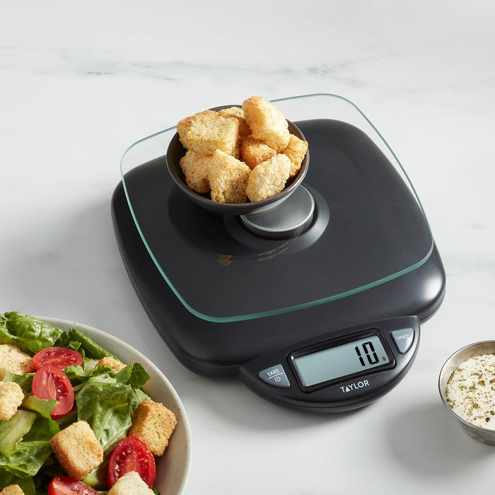 Digital kitchen scale with glass weighing surface holding a bowl of croutons over a salad