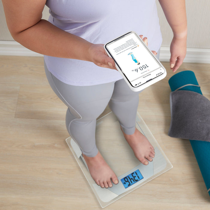 Person stands on a smart weight scale while a phone shows the Precision Hub app interface.