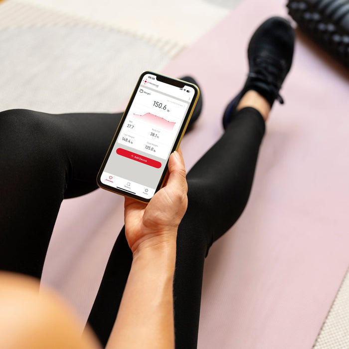 Phone displaying weight tracking app with weight reading and graph, held over a person's leg.