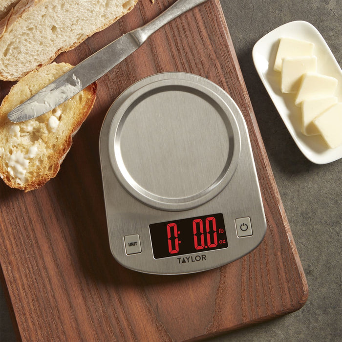Top-down view of a stainless steel digital kitchen scale on a wooden board with bread, butter, and a knife nearby.