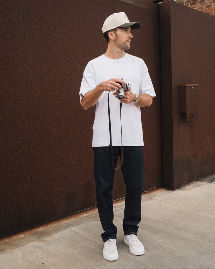 A man stands outdoors holding a camera, wearing The Jeans Relaxed Straight Fit in rinse wash, paired with a white t-shirt, white sneakers, and a cap by c'est normal.