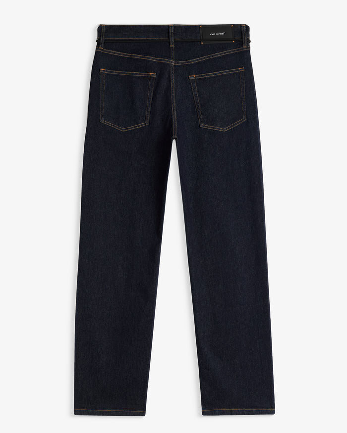 A back view of The Jeans Relaxed Straight Fit in Rinse Wash, laid flat on a white background. The image highlights the straight leg silhouette, two back pockets, subtle orange accent stitching, and the rubber patch at the back waist with the c'est normal logo.