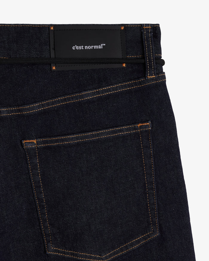 A close-up shot of the back of The Jeans Relaxed Straight Fit in Rinse Wash, laid flat on a white background. The image highlights the back pocket, subtle orange accent stitching, and the black rubber patch at the back waist featuring the c'est normal logo.