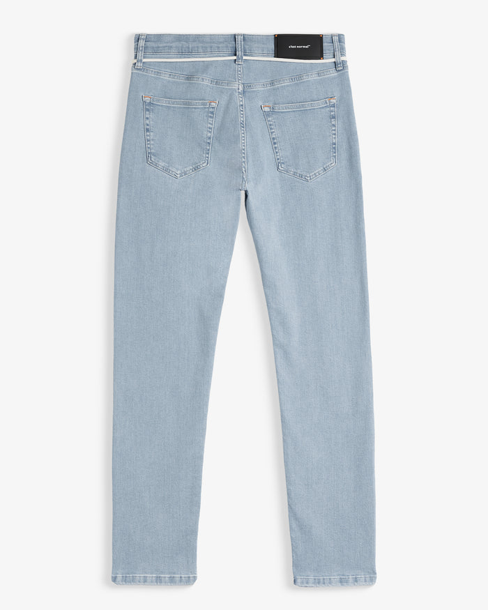 A back view of The Jeans Skinny Fit in light blue, laid flat on a white background. The image highlights the two back pockets, subtle orange accent stitching, and the black rubber patch at the back waist with the c'est normal logo.