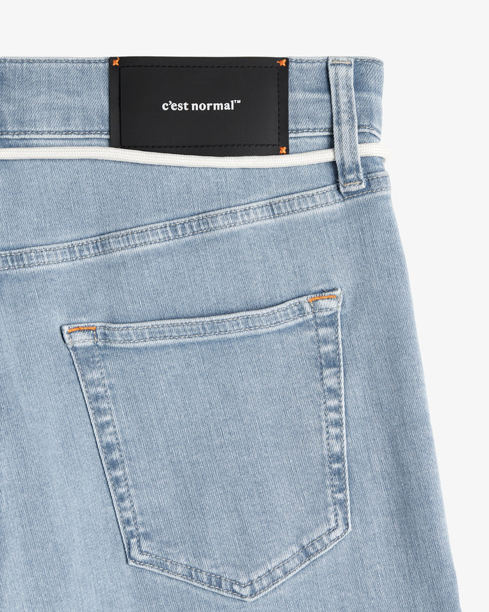 A close-up shot of the back of The Jeans Skinny Fit in Light Blue. The image highlights the black rubber patch with the c'est normal logo at the back waist, subtle orange accent stitching, and the back pocket details.