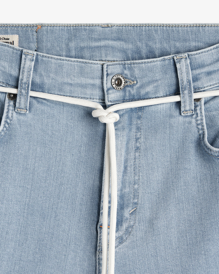 A close-up view of the front waistband of The Jeans Skinny Fit in light blue, featuring a white drawstring tied at the center and a branded c'est normal button.