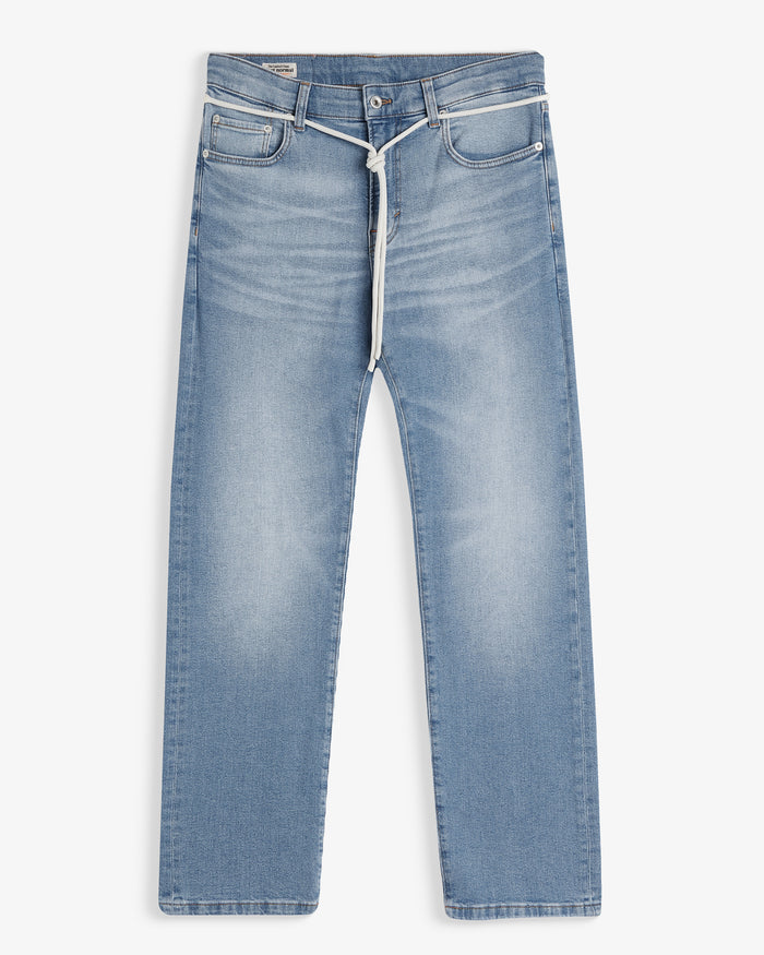 A front view of The Jeans Slim Fit in Light Midwash, laid flat on a white background. The image highlights the clean slim leg silhouette, subtle fade patterns, front pockets, zip fly with top button, and a white drawstring tied around the waist.