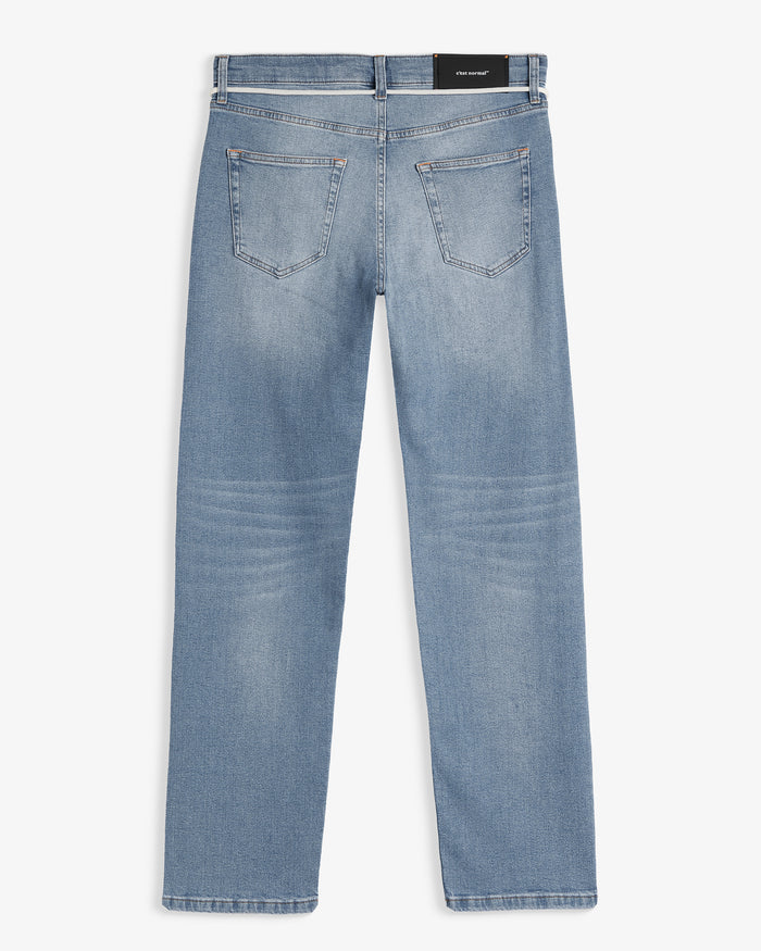 A back view of The Jeans Slim Fit in Light Midwash, laid flat on a white background. The image highlights the two back pockets and the black rubber patch at the back waist with the c'est normal logo.