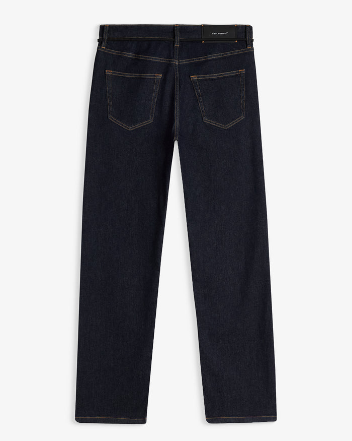 A back view of The Jeans Slim Fit in Rinse Wash, laid flat on a white background. The image highlights the dark denim fabric, orange accent stitching on the pockets and seams, and the black rubber patch at the back waist with the white c'est normal logo.