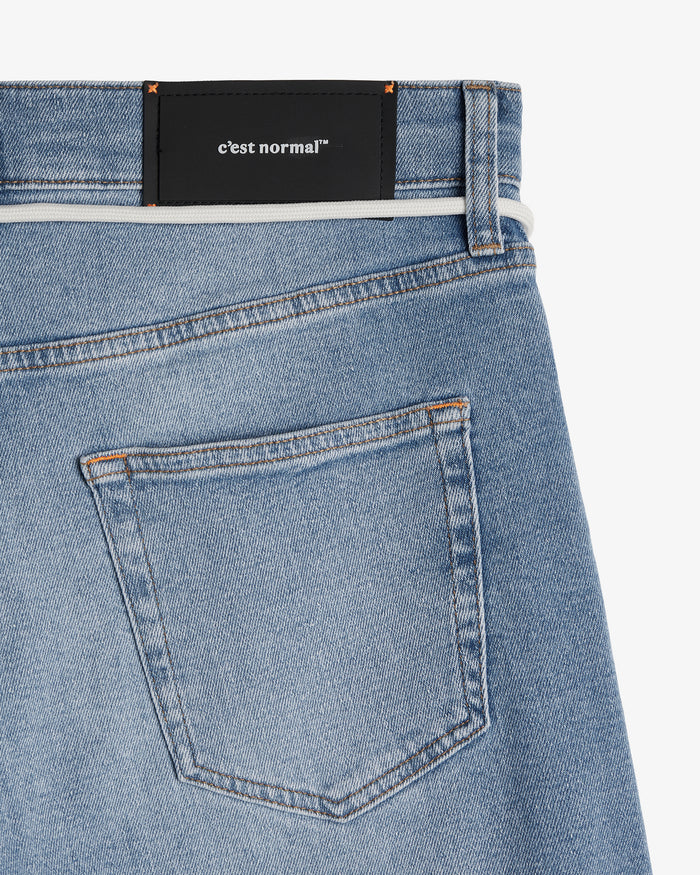 A close-up view of the back of The Jeans Slim Fit in Light Midwash, highlighting the back pocket, the black rubber patch at the back waist with the c'est normal logo, and the subtle orange accent stitching.