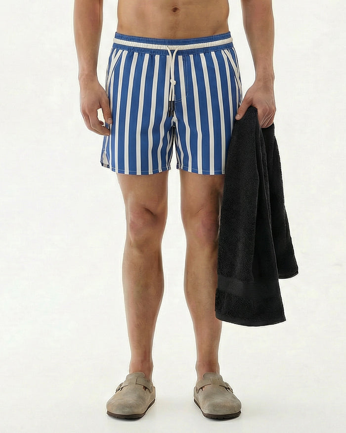 A model wearing The Shorts in Nautical Stripe, featuring a blue and white striped pattern, holding a black towel. The image showcases the shorts with their elastic waistband and drawstring detail, perfect for training or swimming by c'est normal.