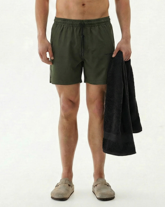 A front view of a model wearing The Shorts in khaki, holding a black towel, standing against a white background. The image highlights the elasticated waistband with a drawstring and the c'est normal logo.