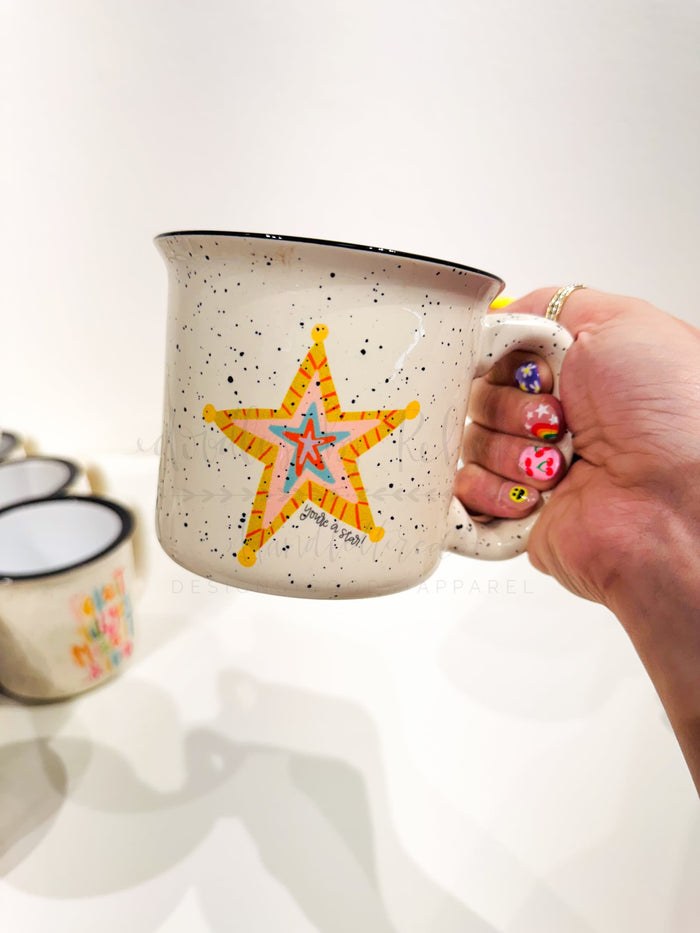 A ceramic mug with a star pattern and speckled background, held in a hand with colorful nail polish.